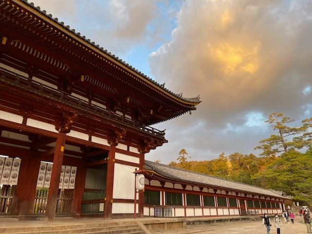 A low-angle view of the large wooden gate and long gallery of Todai-ji temple in Nara, with warm golden clouds and autumn-colored trees filling the sky above.