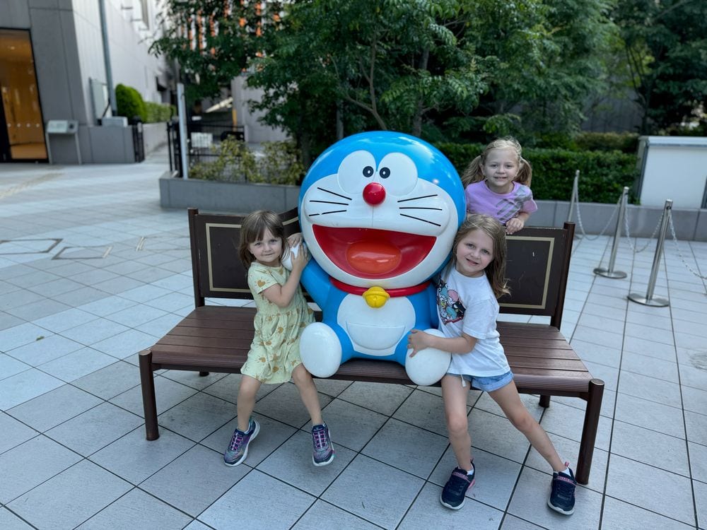 Two young girls stand next to a life-sized Doraemon character statue seated on a bench outdoors at DiverCity Tokyo Plaza in Odaiba.