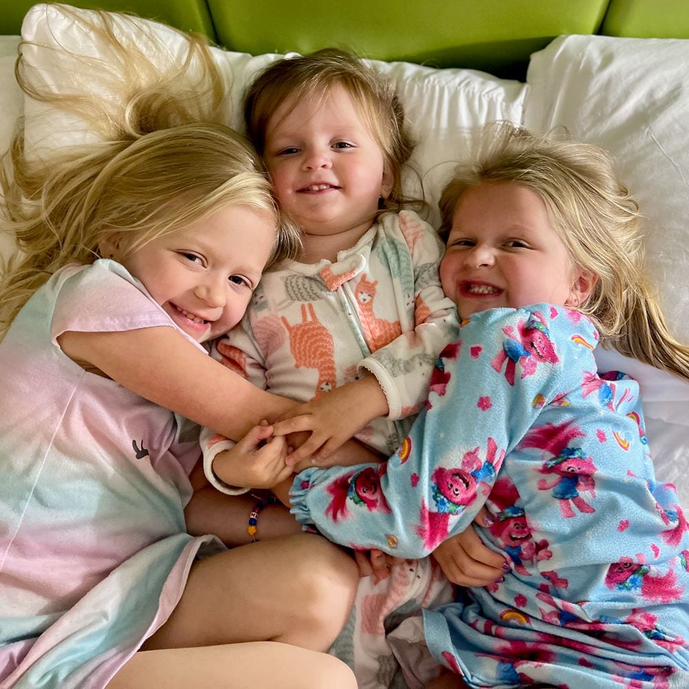 Cora, Harper, and Lily in pajamas piled together on a hotel bed in Brooklyn the night before boarding