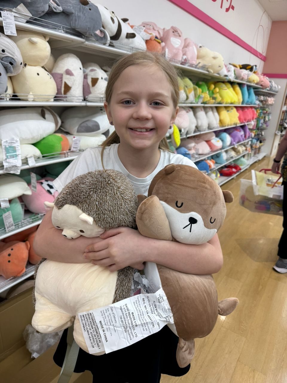 A young girl stands in a colorful Japanese toy shop holding two large stuffed animals, a hedgehog and an otter, while shelves of plush toys line the wall behind her.