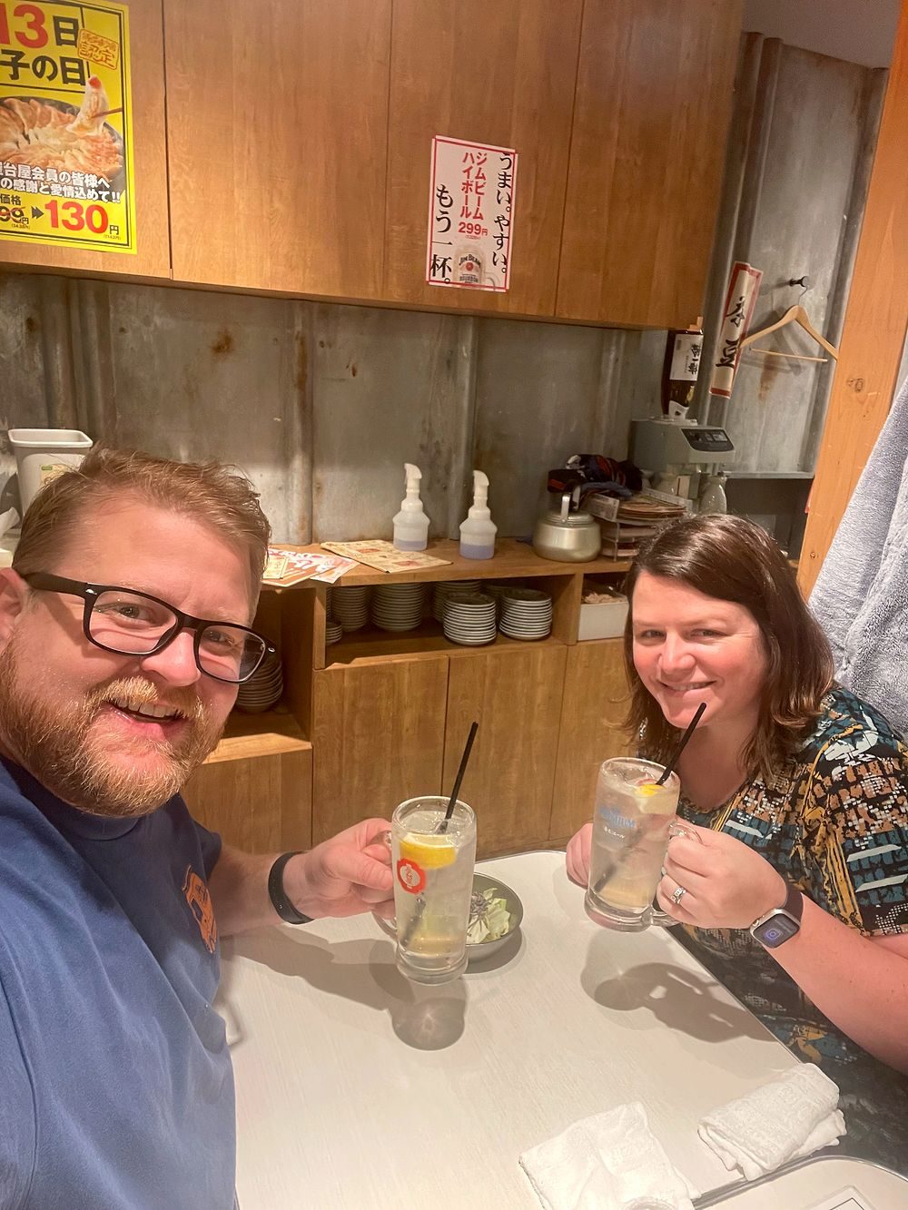 Two people hold up drinks at a small table inside a narrow Golden Gai bar in Shinjuku, Tokyo, with shelves and bottles on the wall behind them.