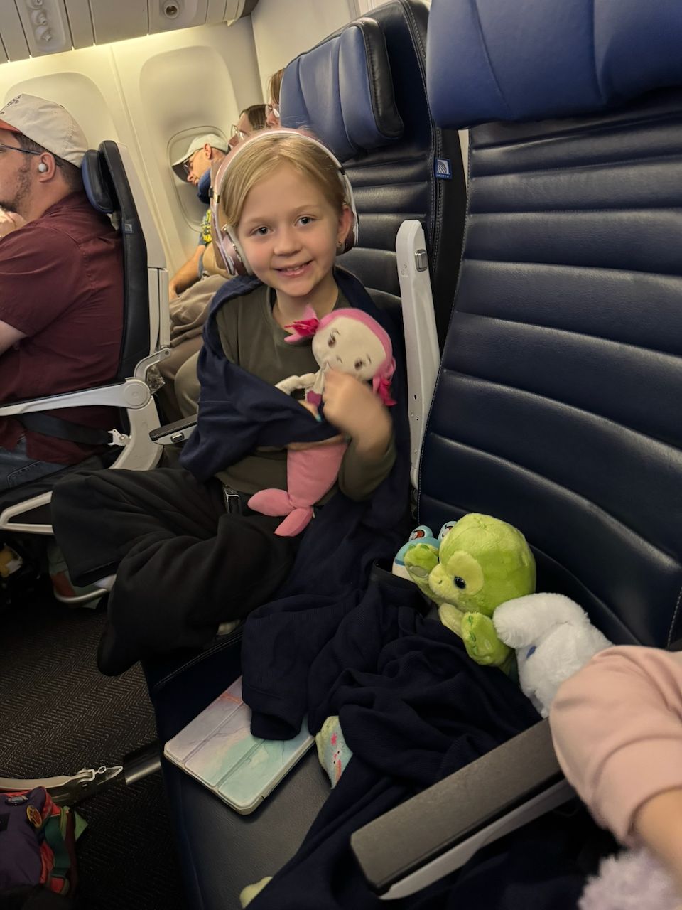 A young girl with pink headphones sits in an airplane seat smiling at the camera, holding a pink and white stuffed doll with a green stuffed turtle beside her.