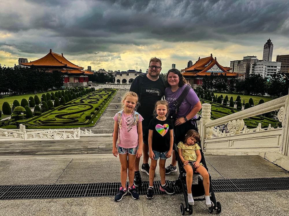 A family of five poses at the top of the steps at Chiang Kai-shek Memorial Hall, with Liberty Square's manicured gardens and a dramatic storm-cloud sky stretching out behind them.
