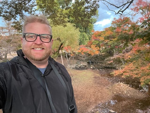 Adam takes a selfie on a path beside a small rocky creek in Nara, Japan, with vivid red and orange autumn maples covering the hillside behind him.