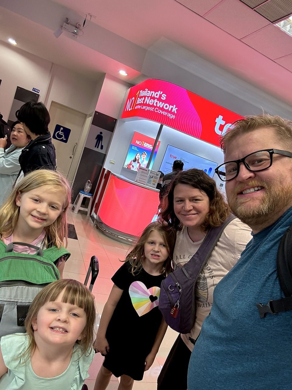 Family of five taking an arrival selfie in Chiang Mai airport terminal in front of a lit True Move network kiosk with their luggage