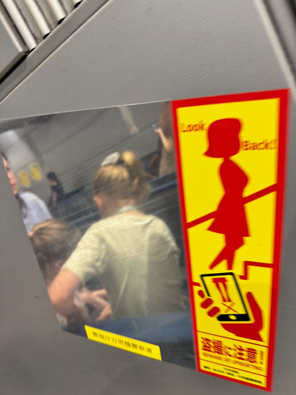 A yellow and red warning sticker on a Japanese subway escalator panel reads Look Back with a pictogram of a woman, reflected in a mirror showing escalator riders behind it.