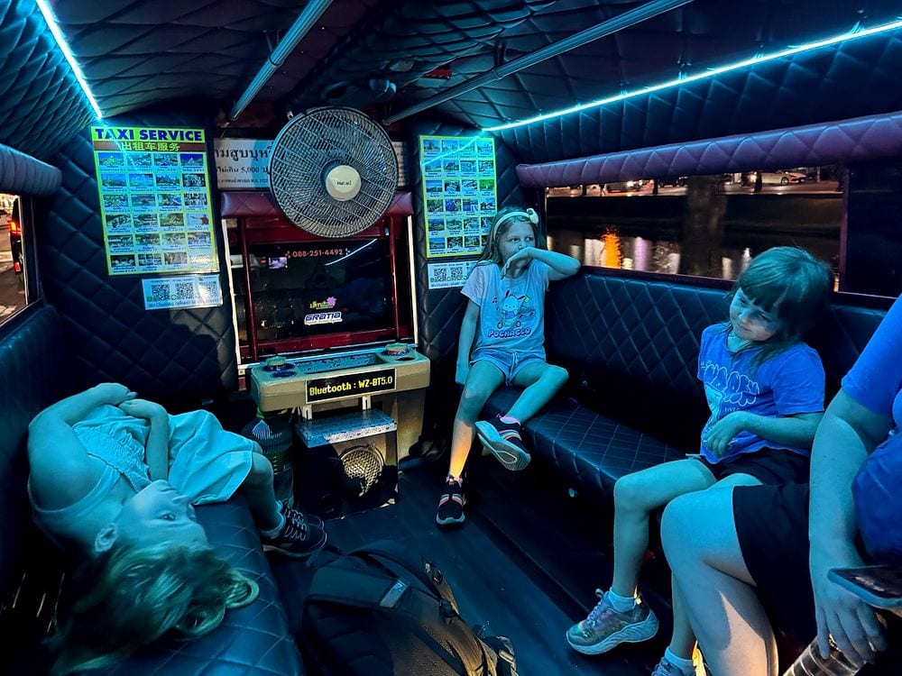 Three girls sit inside a tuk tuk at night in Chiang Mai, lit by blue LED strips, with one girl lying down across the seat and two others sitting across from her looking tired.