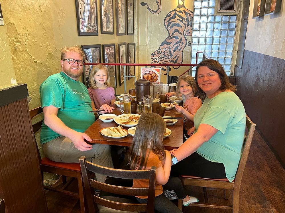 A family sits at a wooden counter inside a retro gyoza restaurant in Asakusa, Tokyo, with a painted tiger mural and framed photos on the wall behind them.