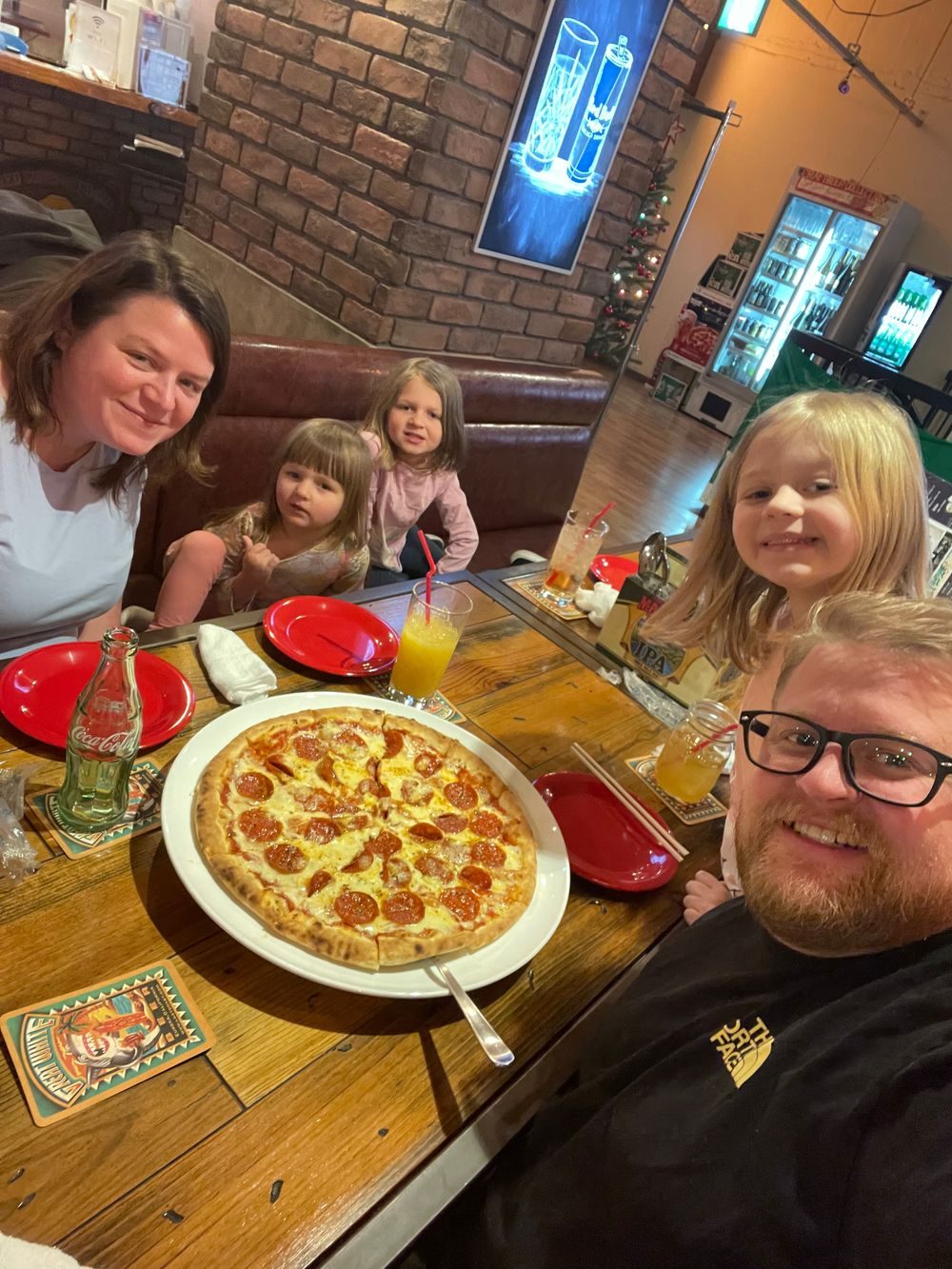A family sits at a wooden table in a casual pizza restaurant in Chiba, Japan, with a pepperoni pizza, drinks, and red plates on the table.