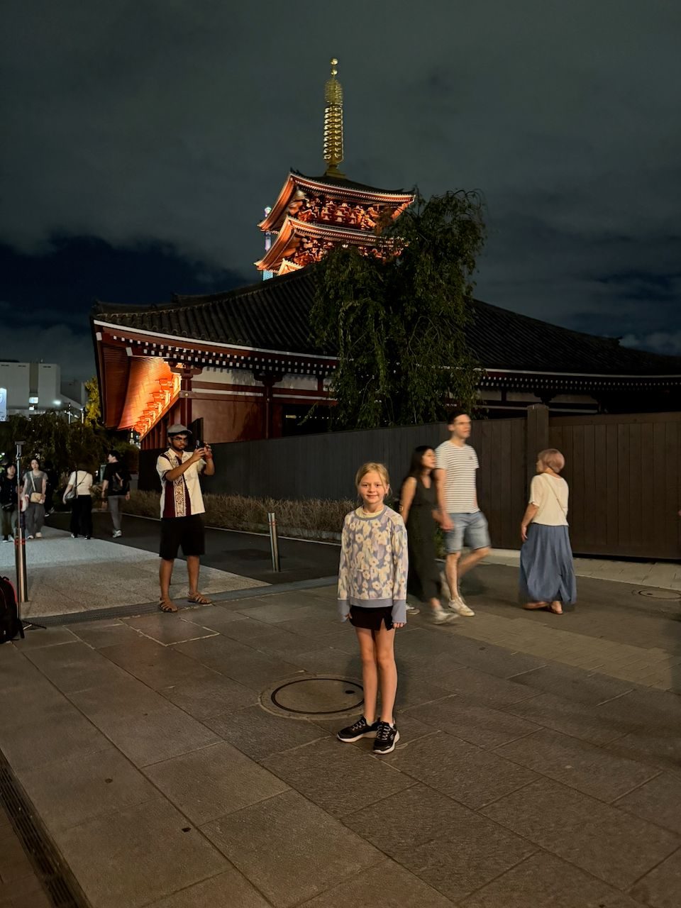 A young girl stands smiling in front of the illuminated five-story pagoda and main hall of Senso-ji temple at night in Asakusa, with other visitors and a man photographing the temple visible in the background.