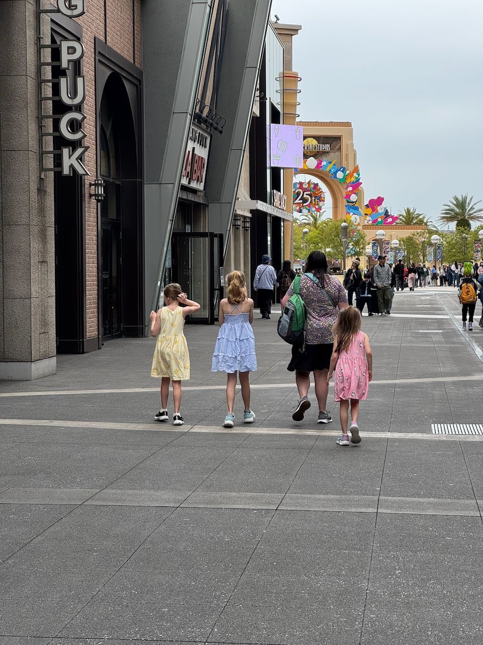 A woman and three girls in summer dresses walk away from the camera toward the Universal Studios Japan entrance arch, with the colorful 25th anniversary decorations visible ahead.