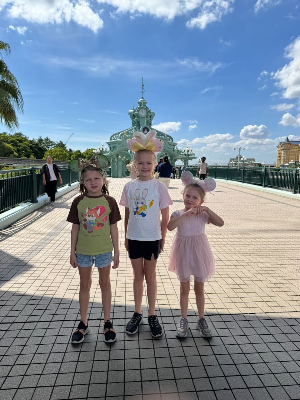 Three girls wearing Mickey Mouse ears stand on the entrance bridge at Tokyo DisneySea with the ornate Mediterranean Harbor building behind them