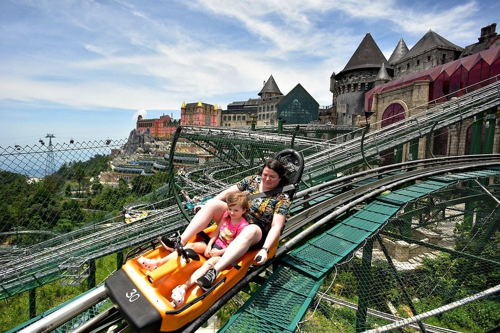 A woman and a young girl ride a yellow alpine coaster cart on a steep metal track at Ba Na Hills, with a sprawling castle complex and mountain scenery visible in the background under a partly cloudy sky.