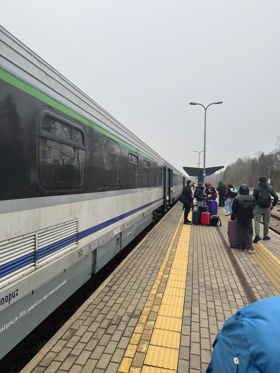 A passenger train at the brick platform in Pagrudai, Lithuania, with travelers and luggage waiting in overcast winter conditions at the border crossing