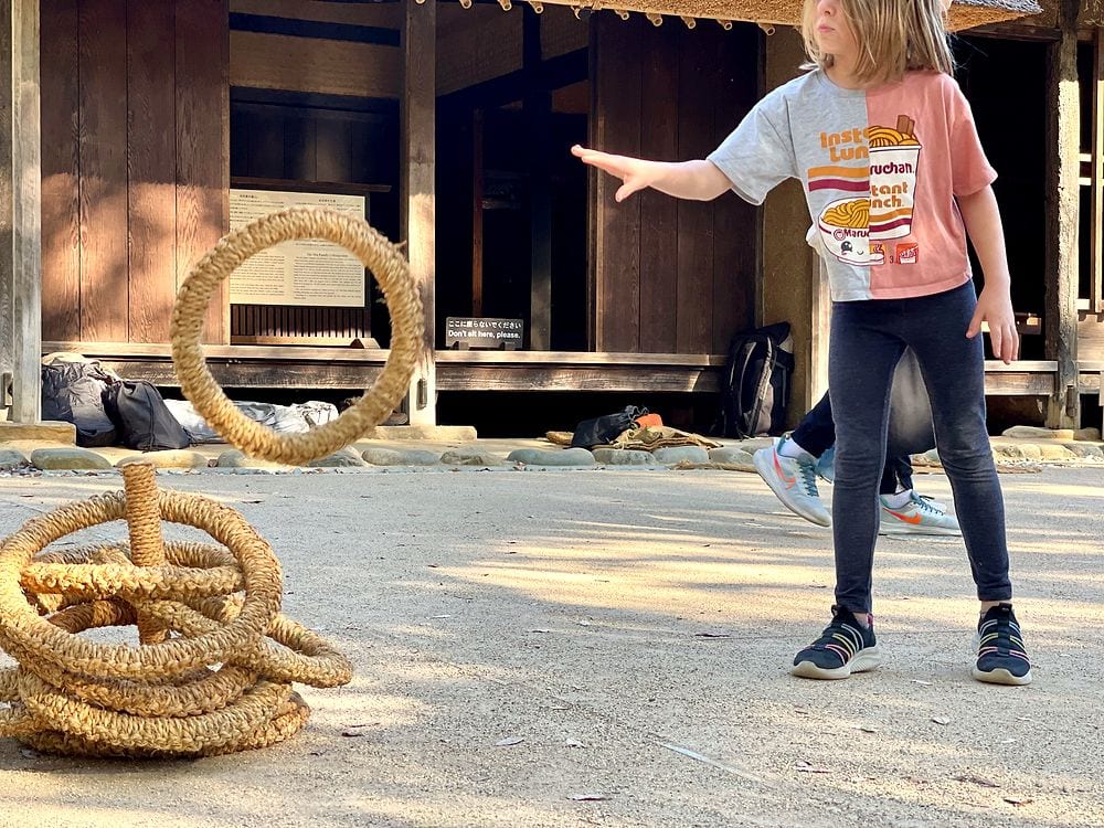 A young girl plays traditional ring toss, tossing a woven straw ring toward a target on the ground in front of a thatched historic building.