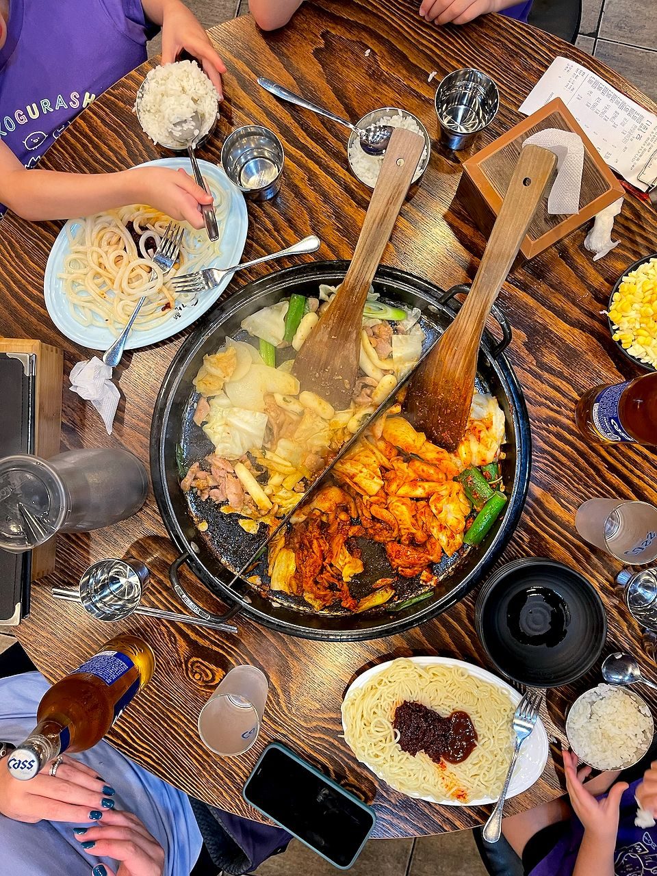 Overhead shot of a divided iron pan with kimchi stir-fry, noodles with gochujang, rice bowls, and a bottle of Cass beer at a Korean restaurant table