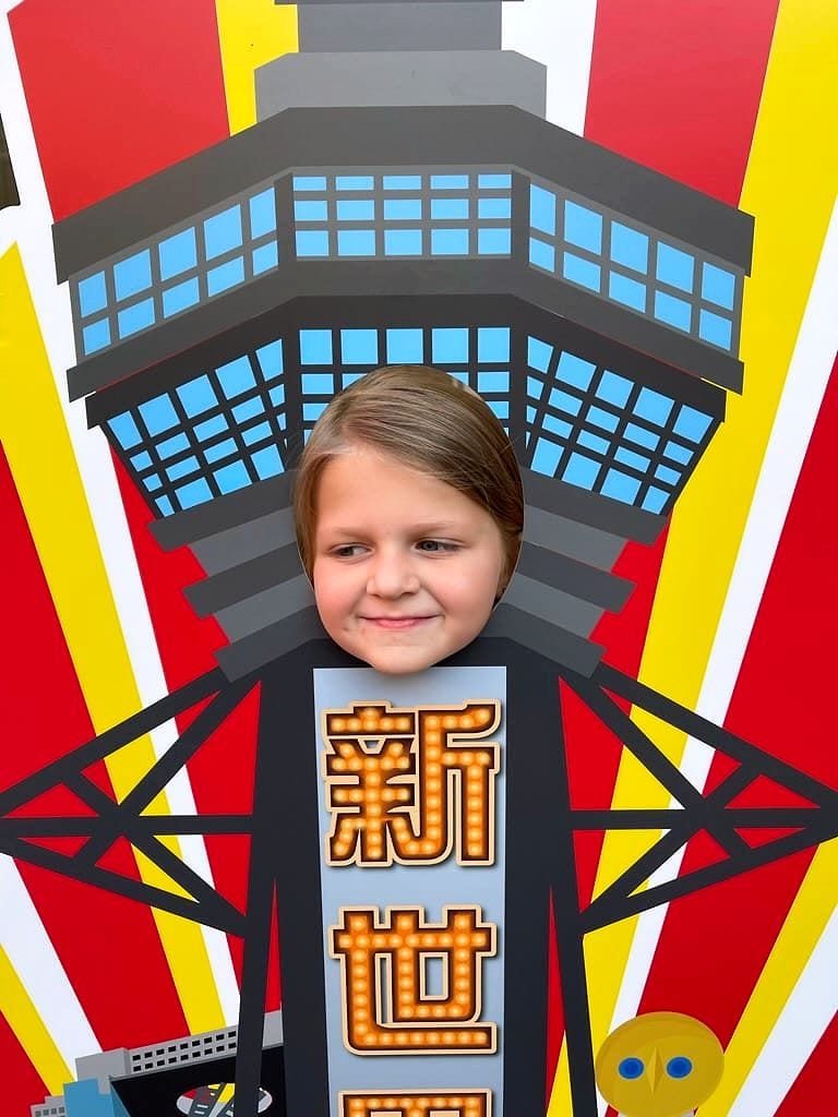 A child's face peeking through a face cutout of Tsutenkaku Tower on a bold red and yellow illustrated Shinsekai-themed board with Japanese kanji characters.