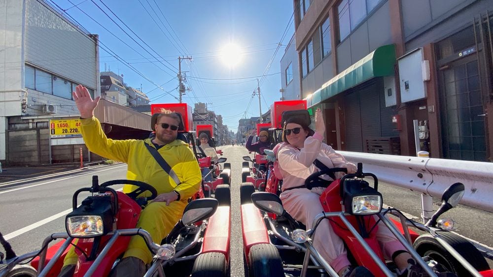 Adam in a yellow Pikachu jumpsuit and Lindsay in a pink My Melody costume drive open-air go-karts side by side down a Tokyo street on a sunny day, both waving at the camera.