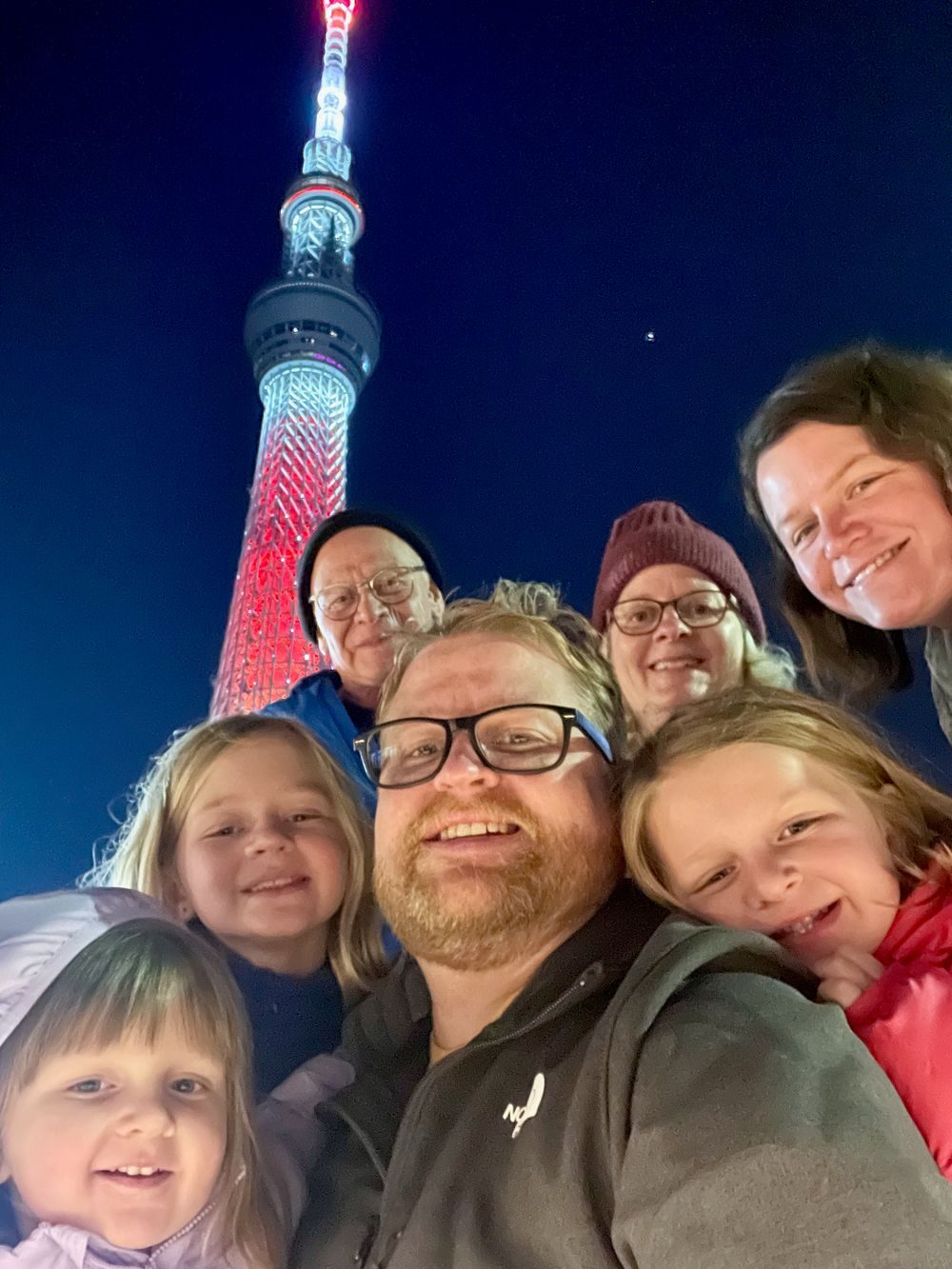 A group selfie of seven people including Adam, Lindsay, three young girls, and two older adults in winter hats, smiling in front of the illuminated Tokyo Skytree at night against a deep blue sky.