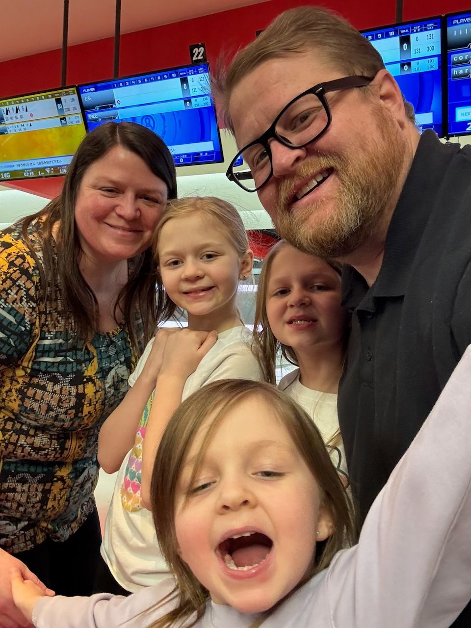 A family selfie at a Japanese bowling alley showing two adults and three girls, with the youngest child in the foreground making an exaggerated open-mouthed expression, Japanese-language scoring screens visible behind them.
