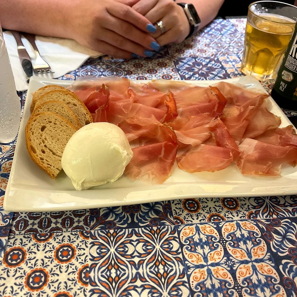 A white rectangular plate of prosciutto, fresh mozzarella, and sliced bread on a blue and orange patterned tablecloth, at a small restaurant in Lucca