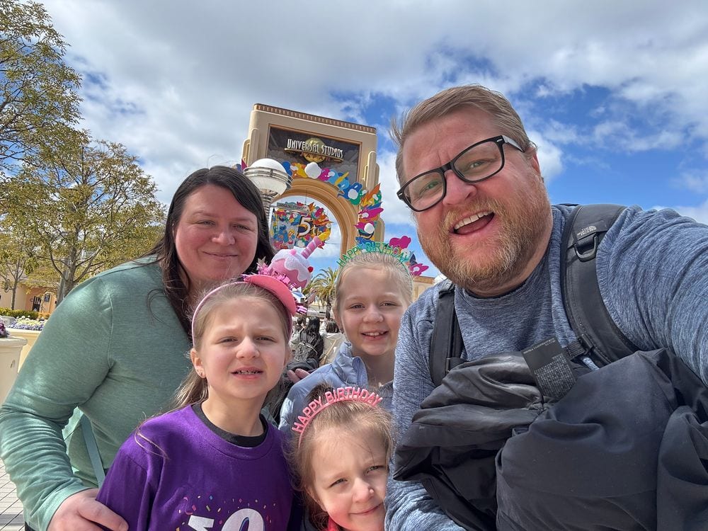 A family selfie in front of the Universal Studios Japan entrance globe, with two girls wearing pink Happy Birthday tiaras under a partly cloudy sky