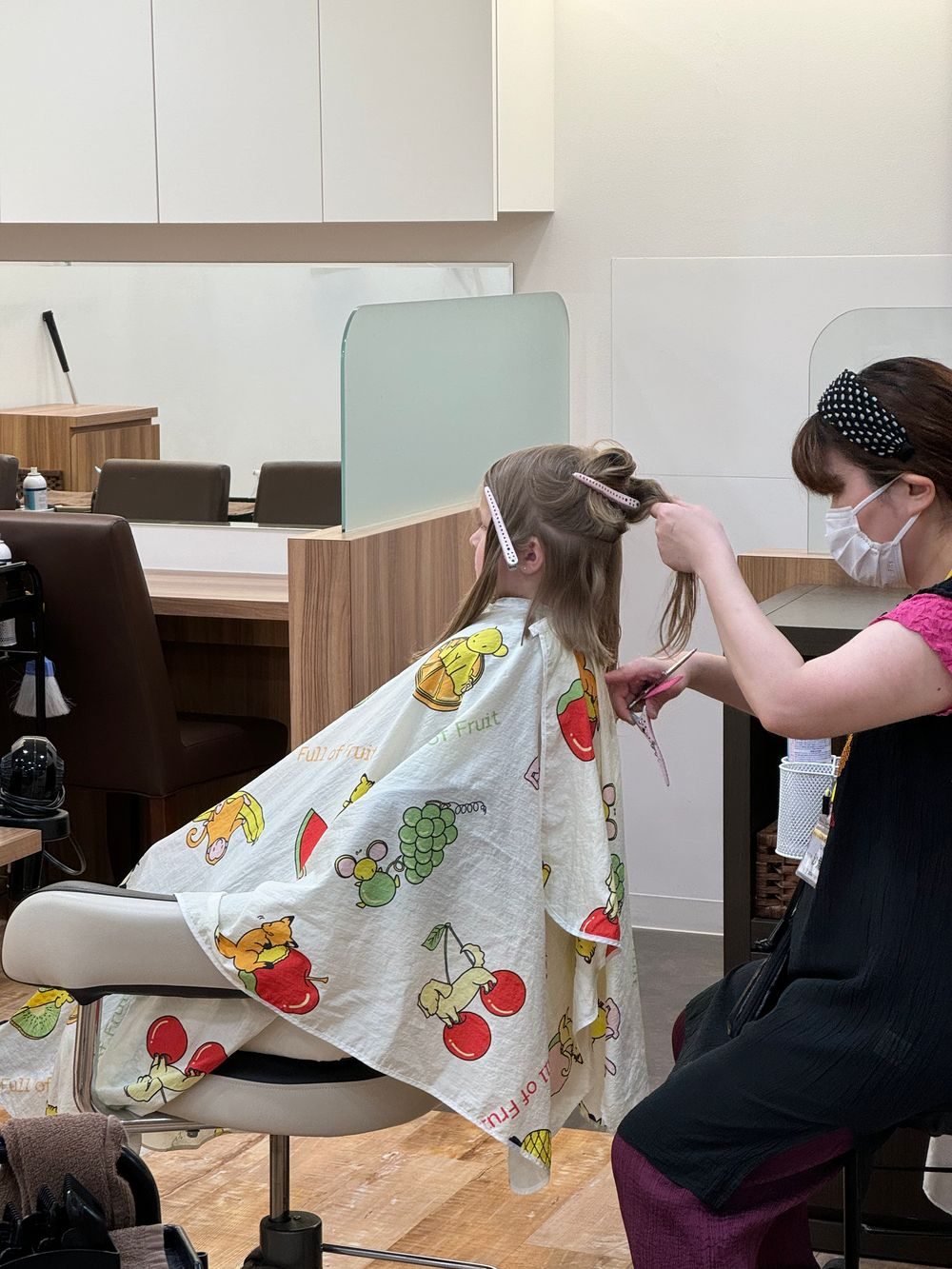 A young girl in pigtails sits in a salon chair wearing a fruit-patterned cutting cape while a masked stylist trims her hair from the side.