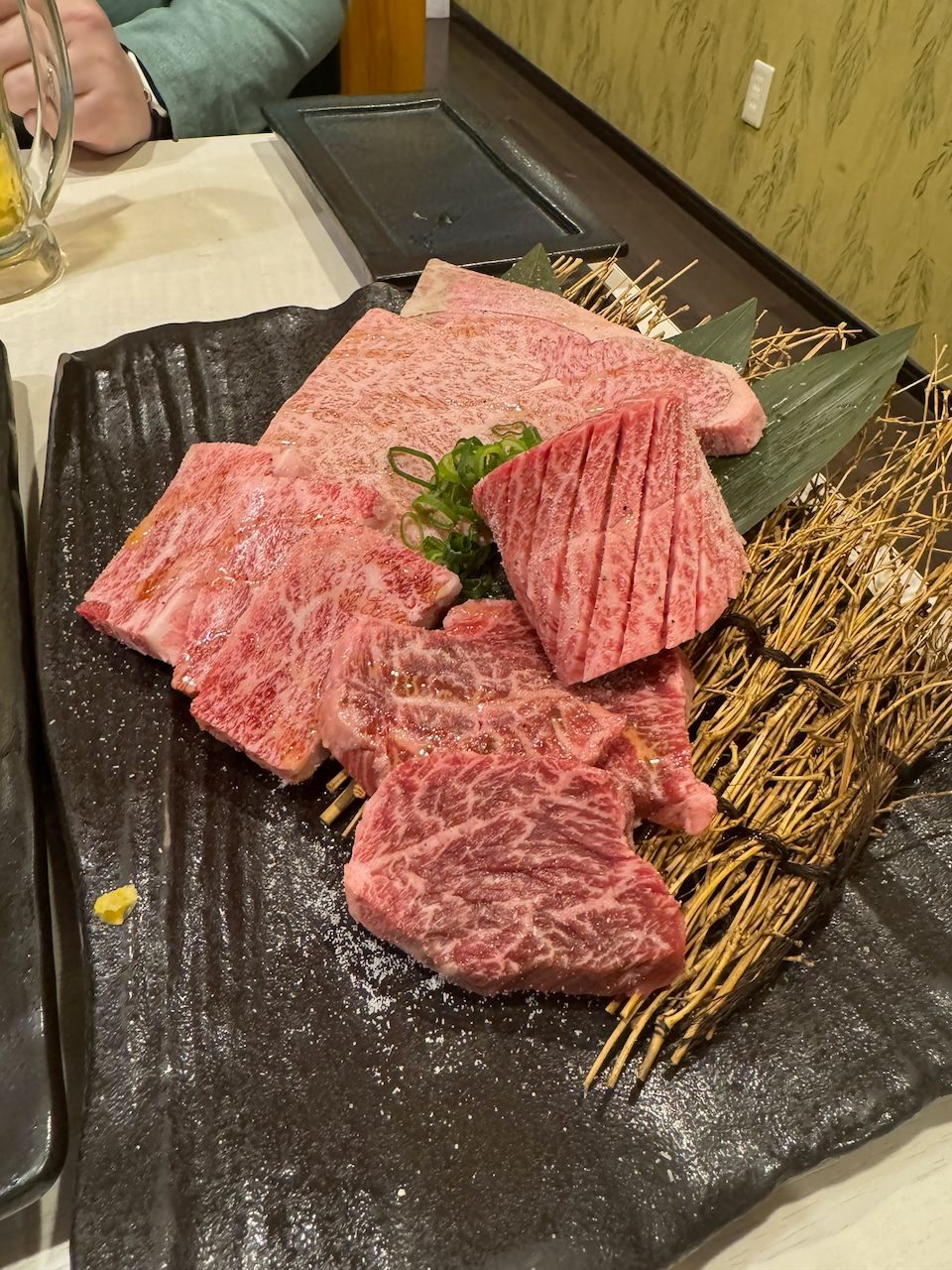 A dark ceramic platter holds multiple cuts of heavily marbled raw wagyu beef arranged with dried straw and green garnish, with a glass of beer visible in the background.