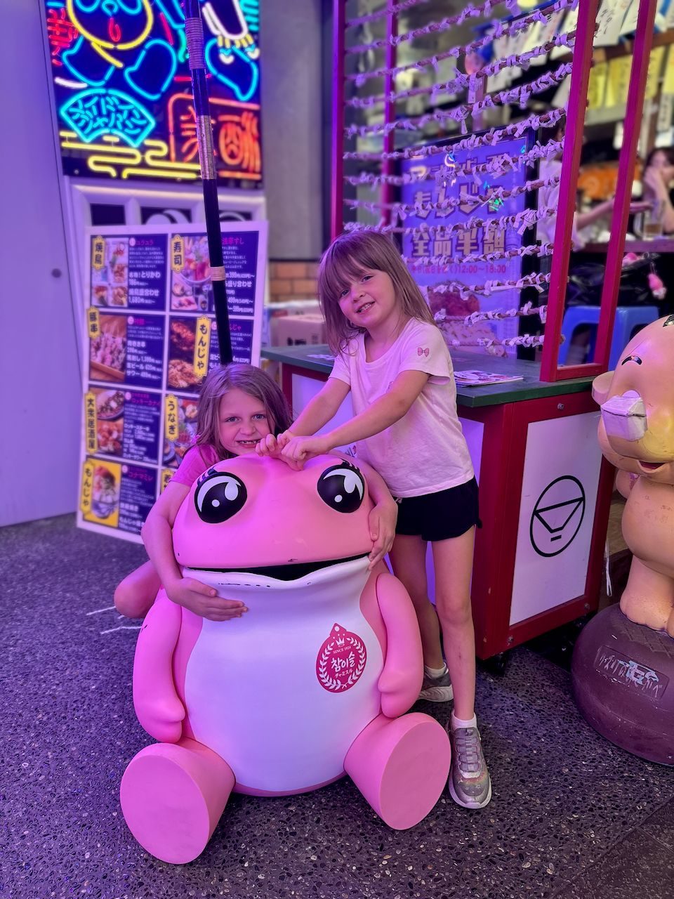 Two young girls pose with a large pink frog mascot statue outside a neon-lit restaurant in Asakusa, Japan, one hugging it from the front and the other standing behind it smiling.