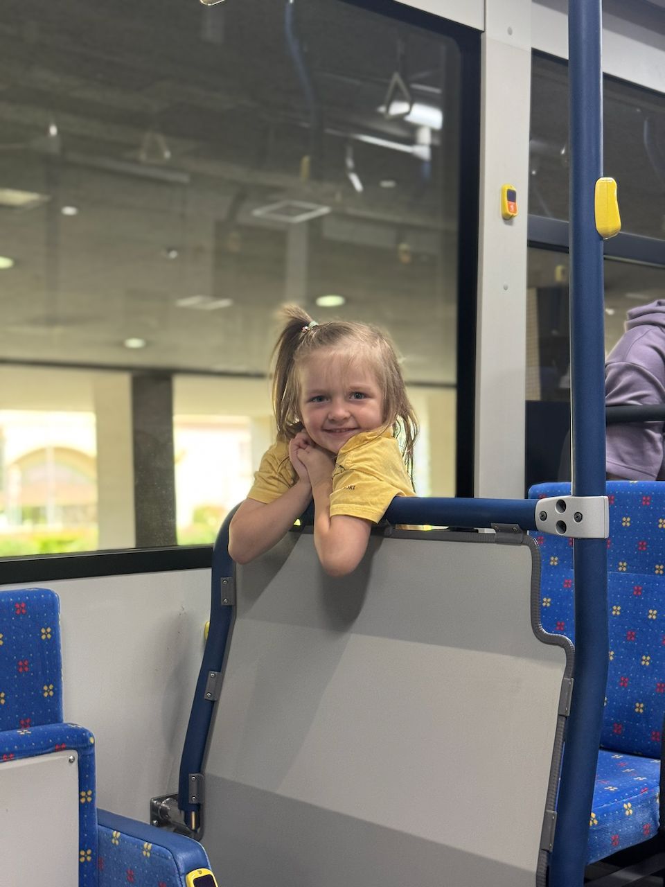 A young girl in a yellow shirt leans over the back of a bus seat with her hands clasped together, smiling at the camera inside a Japanese public bus with blue floral-patterned seats.