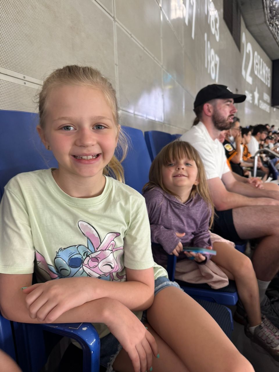 Two young girls sit in blue stadium seats at Tokyo Dome, the older one in a Stitch t-shirt smiling at the camera and the younger one holding a small toy behind her.