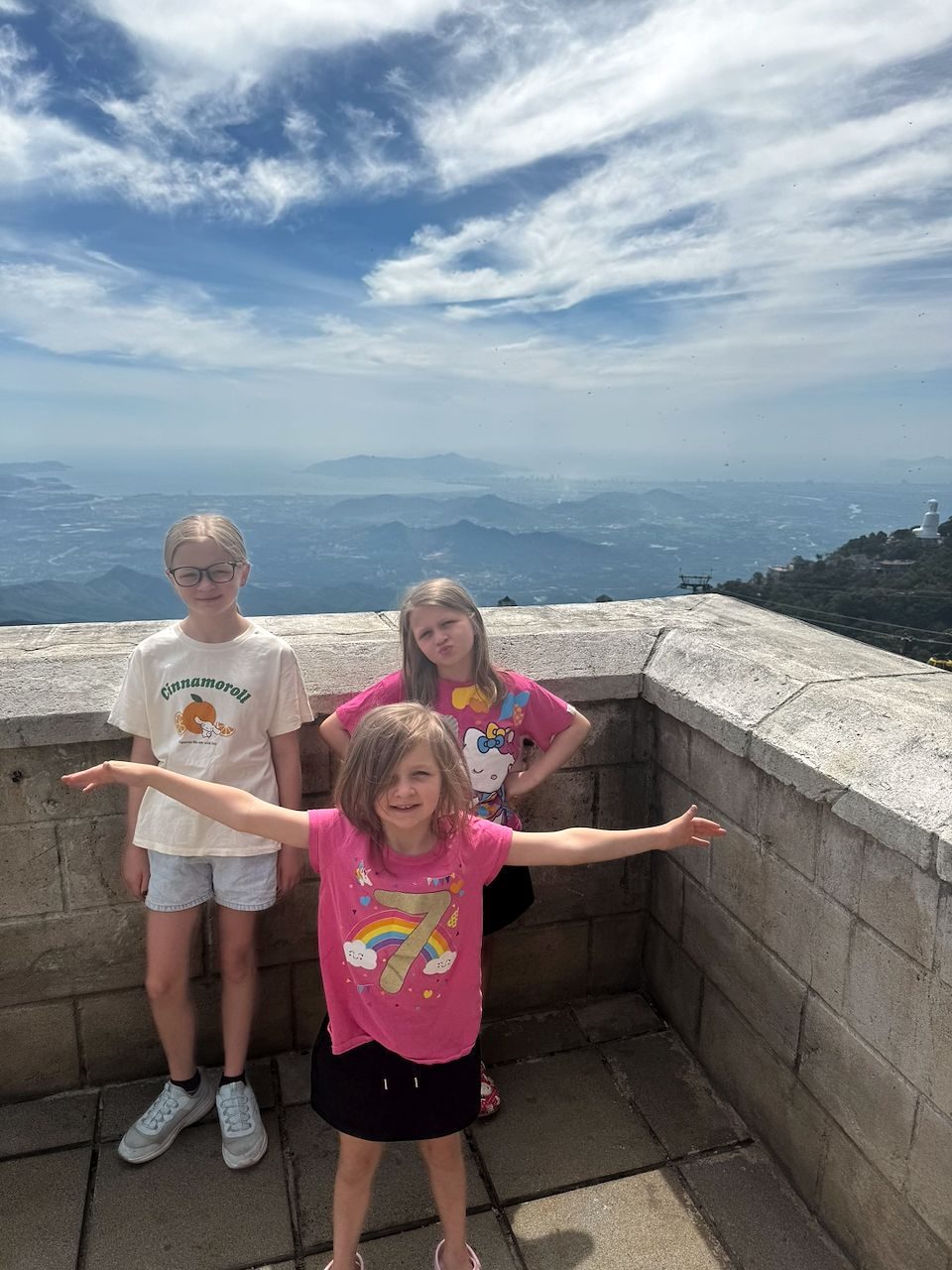 Three girls pose on a stone parapet at the top of Ba Na Hills, with a sweeping hazy mountain and coastal view behind them; the youngest in a pink birthday shirt spreads her arms wide while the older two stand on either side.