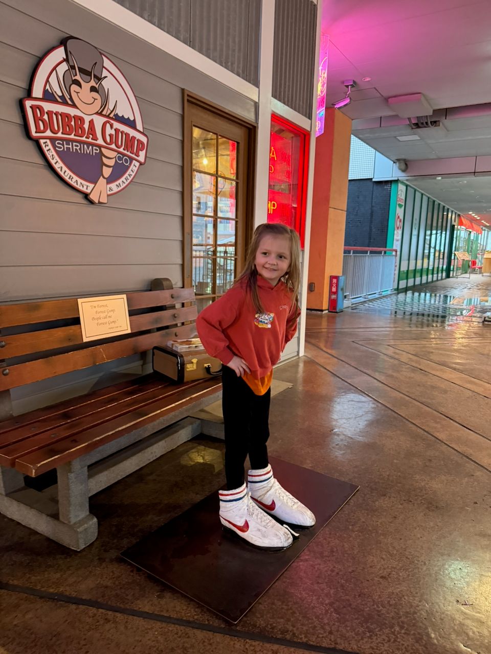 A young girl in a red hoodie and Nike sneakers stands on a Forrest Gump shoe display outside a Bubba Gump Shrimp Co. restaurant on a rainy evening in Osaka, with a wooden bench and the restaurant's sign visible behind her.