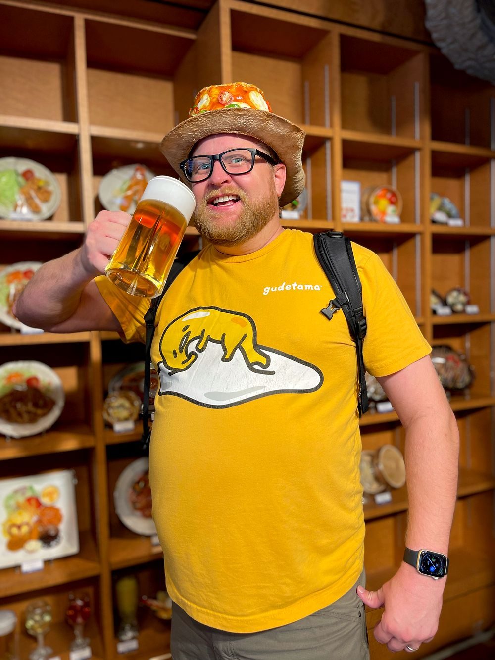 A man wearing a hat shaped like a slice of pizza toast holds a giant mug of beer while posing in a Kappabashi plastic food sample shop in Tokyo.