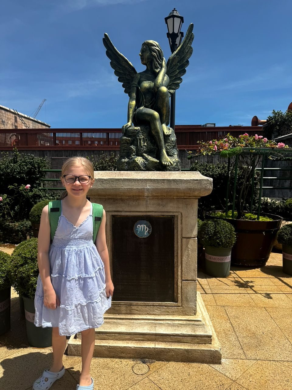 A young girl with glasses and a green backpack stands smiling in front of a bronze winged Virgo zodiac statue on a stone pedestal in a garden courtyard under a blue sky.