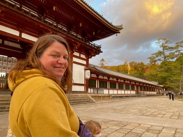 Lindsay, wearing a yellow hoodie and carrying a small child in purple, looks back at the camera in front of the large red wooden gate of Todai-ji temple in Nara, with autumn trees and a dramatic cloudy sky behind her.