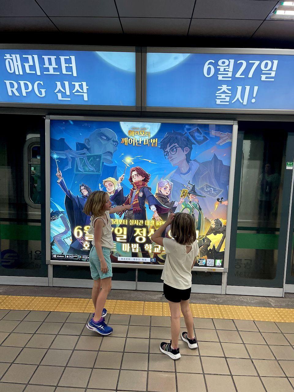 Two children on a Seoul subway platform pointing at a large illuminated Harry Potter game advertisement