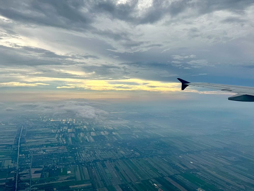 View from an airplane window of Bangkok's city grid and farmland on descent under a hazy sky, with the wing on the right