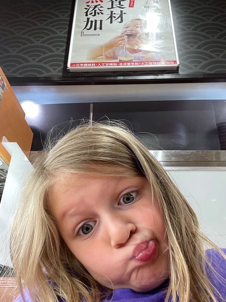 Cora making an exaggerated fish face at the camera inside a small Osaka ramen shop, a Japanese food-quality sign visible on the wall behind her.