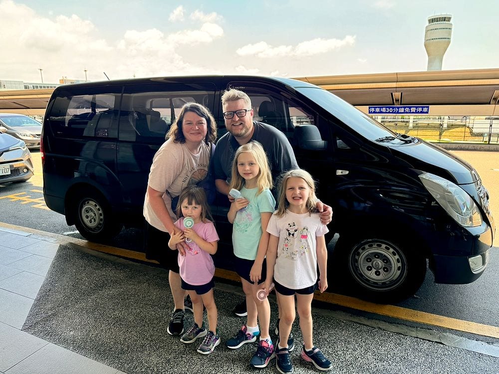 A family of five poses in front of a black van outside Taoyuan International Airport with the air traffic control tower visible in the background.