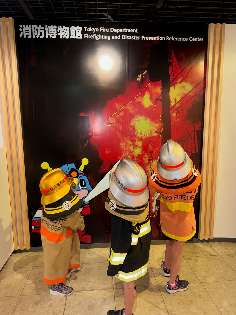 Three young children in firefighter costumes and helmets look at a red and black exhibit display at the Tokyo Fire Museum.