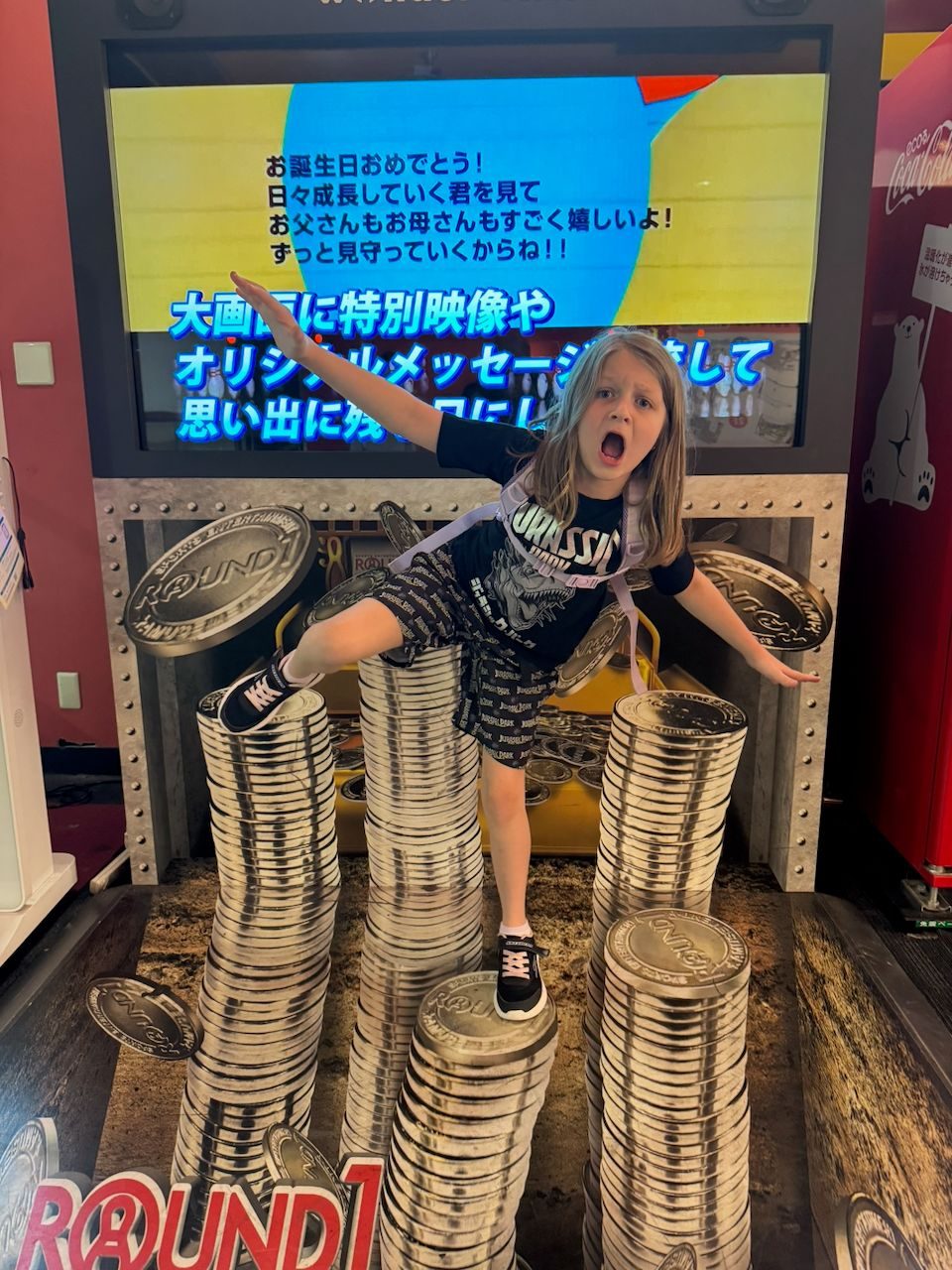 A young girl in a Jurassic Park t-shirt stands on a giant stacked-coins photo op installation at Round1 entertainment center in Japan, arms spread wide and mouth open in an exaggerated roar, with a Japanese birthday message displayed on a screen behind her.