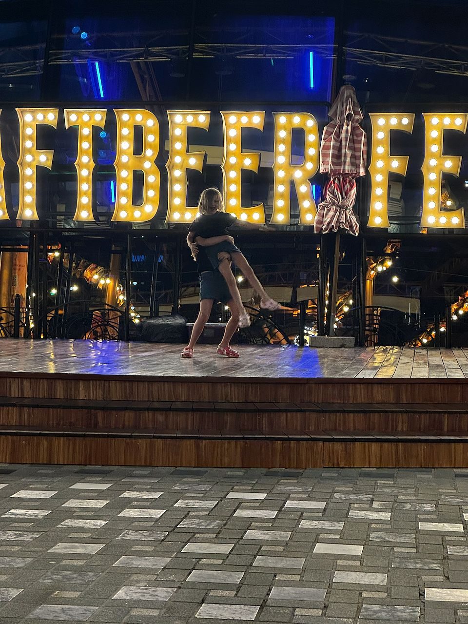 Two children dancing together on a wooden stage in front of large illuminated marquee letters spelling Craft Beer Fest at an outdoor venue at night.