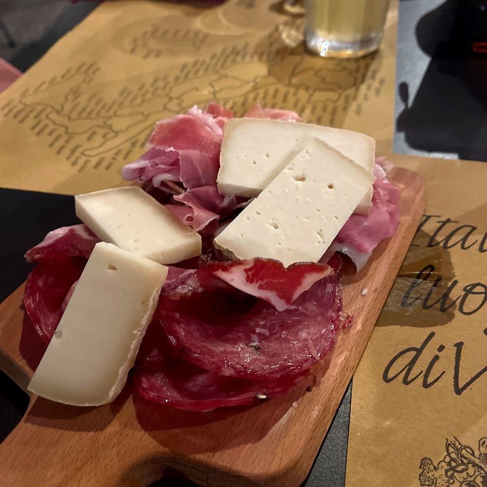 A wooden cutting board covered with sliced cured meats including salami and prosciutto, topped with wedges of white cheese, at a restaurant in Lucca