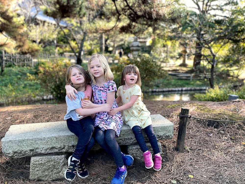 Three girls sit together on a stone bench in a Japanese garden, pine trees and greenery behind them.