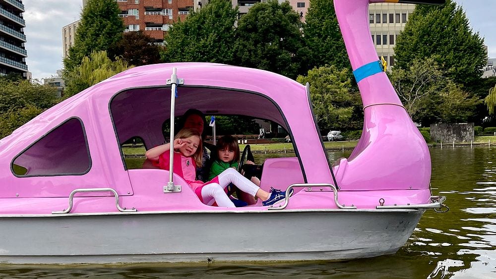 Two young girls ride together in a pink swan-shaped paddle boat on Shinobazu Pond in Ueno Park, Tokyo, with city buildings visible across the water.