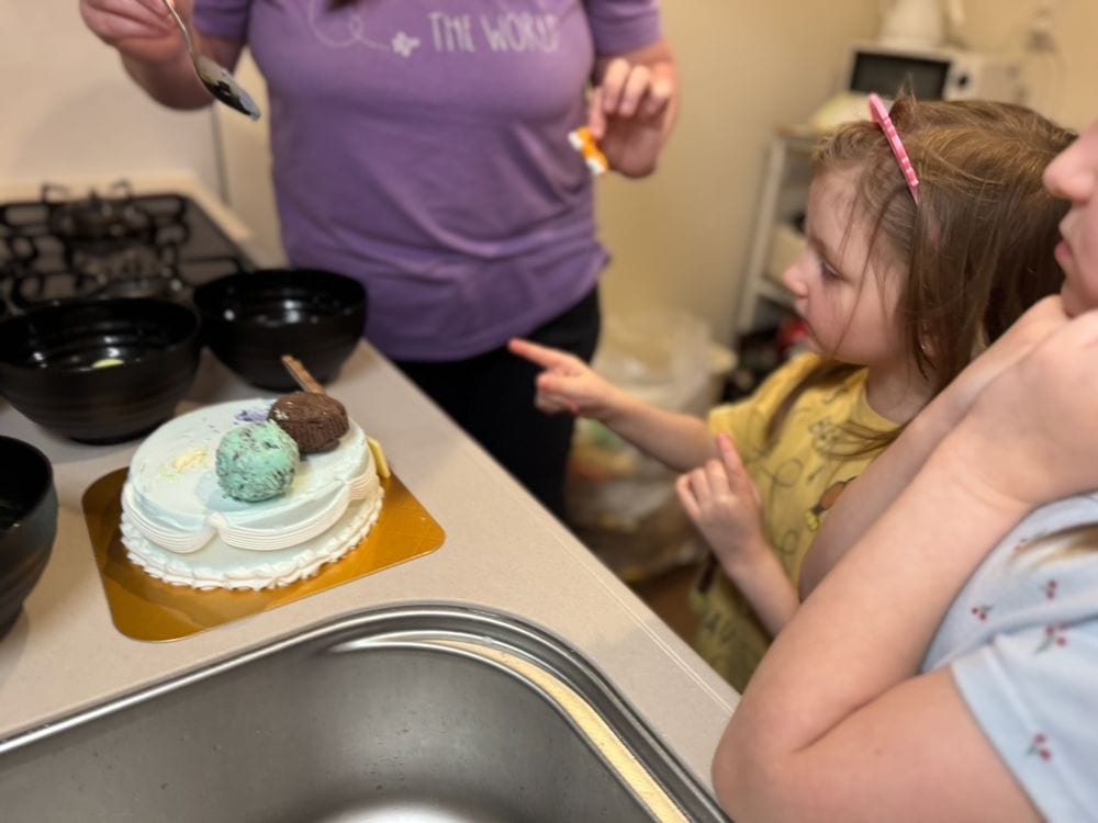 A young girl with a pink headband points at a white cake topped with two ice cream scoops on a kitchen counter in an Osaka apartment, with an adult standing behind the counter holding a spoon.