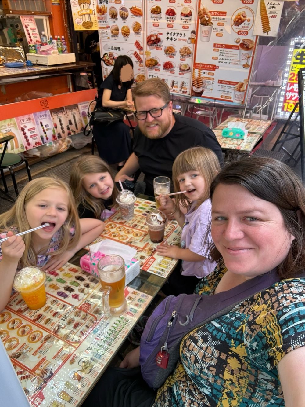 A family of five sits at an outdoor food stall counter in Ameyoko, Ueno, Tokyo, with menu photos lining the counter in front of them.