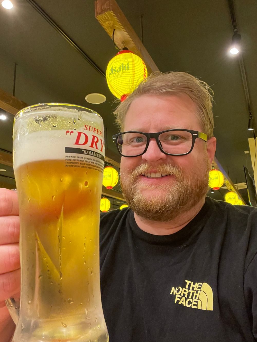 A man holds up a glass of draft beer at an izakaya in Chiba, Japan, with red and yellow Asahi-branded lanterns hanging overhead.