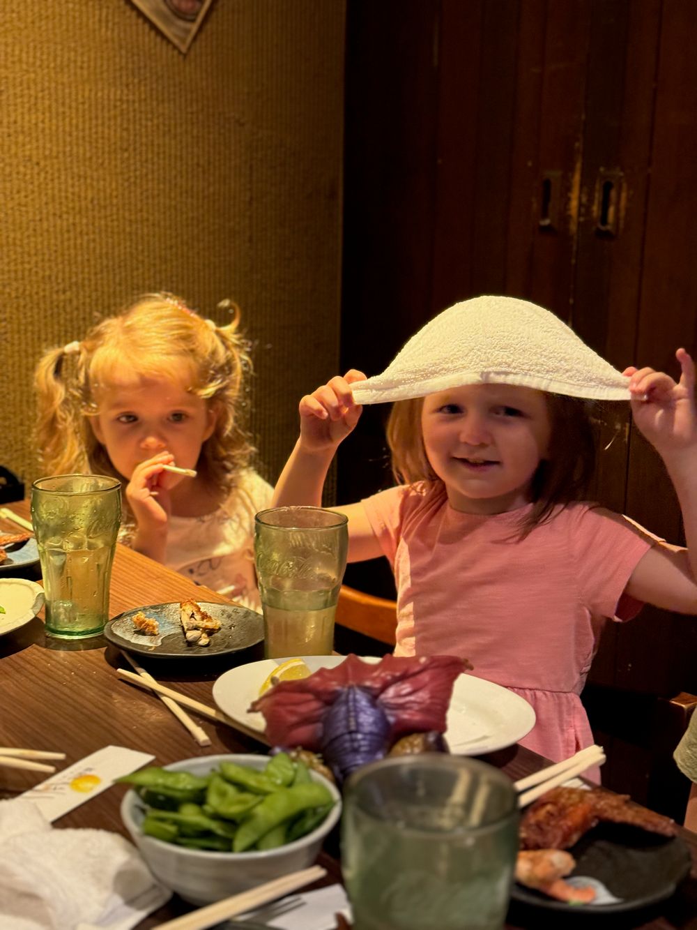 Two young girls sit at an izakaya table in Tokyo, one holding up a white hand towel, with a reddish crab leg visible on a plate among other dishes.